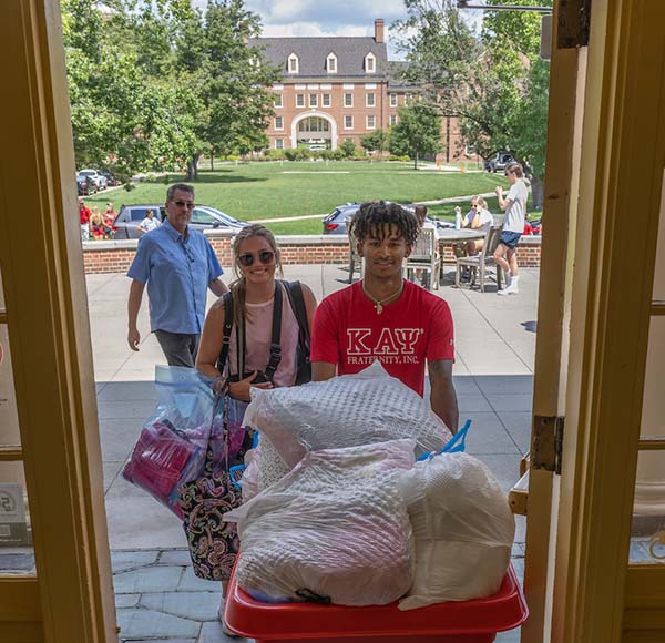 A student, framed by a doorway in tappan hall, pushes a red bin full of move-in dorm room items. Morris hall is in the background